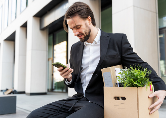 Man sat with office supplied in box, looking at smartphone